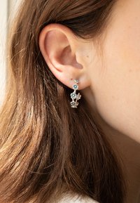 Orecchini d'argento con fiori blu, design delicato con accenti floreali, indossati sull'orecchio, vista ravvicinata con capelli castani visibili.