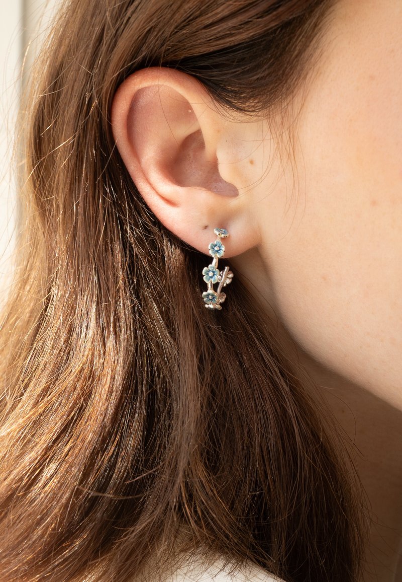 Orecchini d'argento con fiori blu, design delicato con accenti floreali, indossati sull'orecchio, vista ravvicinata con capelli castani visibili.