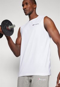 Witte mouwloze t-shirt, van katoen, met ronde hals, voorzien van een rood en marineblauw logo op de borst. Model houdt een zwarte dumbbell vast. Grijze joggingbroek zichtbaar.