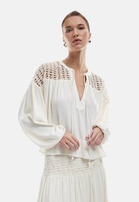 Blusa bianca con scollatura a V, dettagli in pizzo sulle spalle e maniche ampie, realizzata in tessuto leggero e testurizzato.