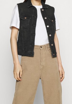Person iført en hvid kortærmet skjorte, sort denimvest med knapper og højtaljede beige kordfløjlsbukser med hænderne hvilende langs siderne.