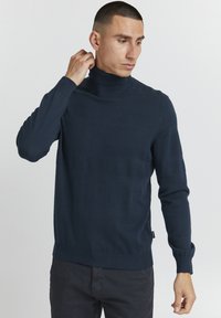 Maglione a collo alto blu navy in un knit testurizzato con sezioni lisce; design aderente, polsini e orlo a coste, e un sottile etichetta con logo sul lato.