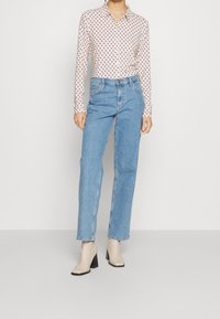 Blusa a maniche lunghe con colletto e un piccolo motivo rosso su uno sfondo chiaro, abbinata a jeans azzurri a gamba dritta e stivali beige alla caviglia.