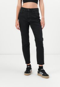 Mujer vistiendo jeans negros de cintura alta y zapatillas Adidas negras con rayas blancas, de pie contra un fondo blanco liso.