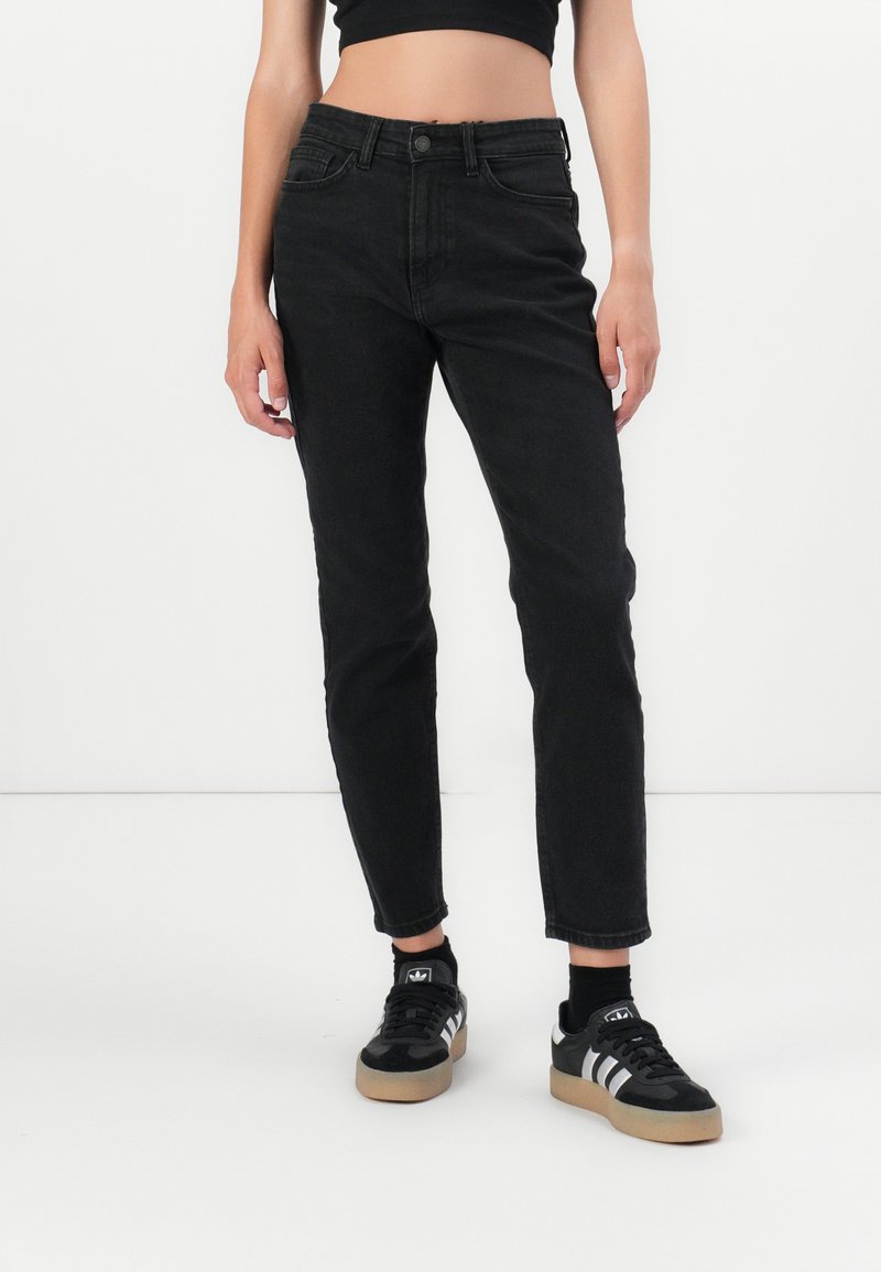 Mujer vistiendo jeans negros de cintura alta y zapatillas Adidas negras con rayas blancas, de pie contra un fondo blanco liso.