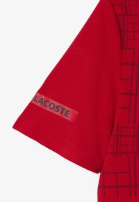 Röd polo t-shirt med ett strukturerat mönster och Lacoste-logotyp på ärmen. Tillverkad av ett mjukt tyg med kort ärm.