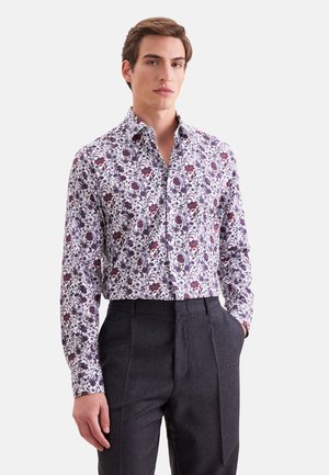 Homme portant une chemise blanche à manches longues avec un motif floral violet et rouge et un pantalon gris foncé, debout avec une main dans la poche.