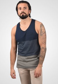 Muškarac s bradom nosi tamnoplavu i sivu majicu bez rukava, bež hlače, te ima detaljnu tetovažu klasičnih figura na desnoj ruci, stoji na jednoličnoj pozadini.