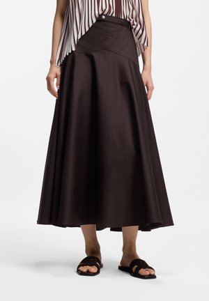 Vrouw met een donkerbruine A-lijn midi-rok met drukknoop aan de voorkant en zwarte open-toe sandalen, staand tegen een witte achtergrond.