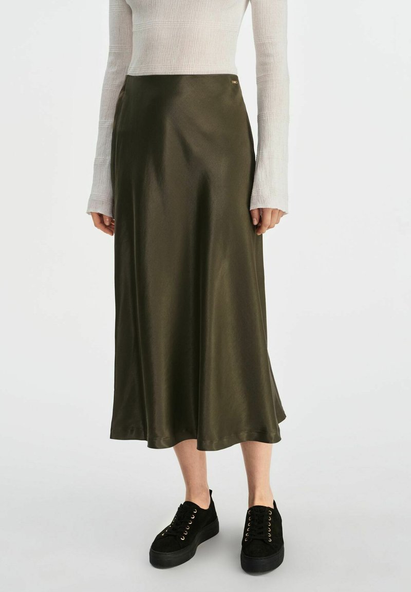 Sukienka midi w oliwkowym satynowym kolorze o gładkiej teksturze, z bocznym rozcięciem i subtelnym metalicznym akcentem. Zestawiona z czarnymi, sznurowanymi trampkami.