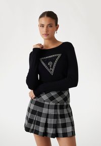 Schwarzer Strickpullover mit weitem Ausschnitt und dreieckigem Logodesign; kombiniert mit einem karierter, gefälteter Minirock in Schwarz und Weiß.