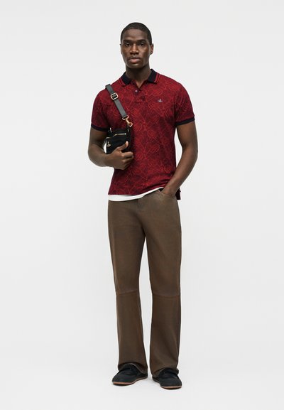 Hombre con camiseta polo de patrón rojo, pantalones marrones, zapatos negros y un bolso bandolera negro sobre un fondo blanco.