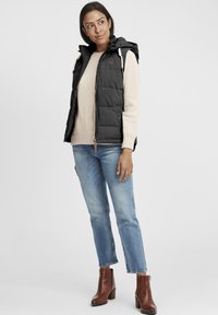 Svart pufferväst med huva, med dragkedjestängning och sidofickor, bärs över en ljusbeige sweatshirts, tillsammans med blåjeans och bruna ankelboots.