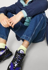 Svart och lila Nike-sneakers, ljusgröna strumpor, blå jeans och en marinblå puffjacka över en teal hoodie, sittande på en grå bakgrund.