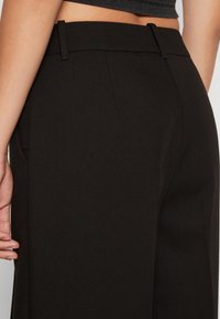 Černé kalhoty na míru s hladkou texturou, výraznými švy a poutky na pásek. Design má vysoký pas a volný střih.