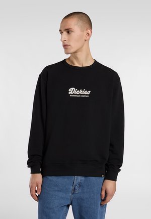 Svart sweatshirt med rundad hals och långa ärmar, med en vitt broderad logga på bröstet. Bärs med blå denimjeans.