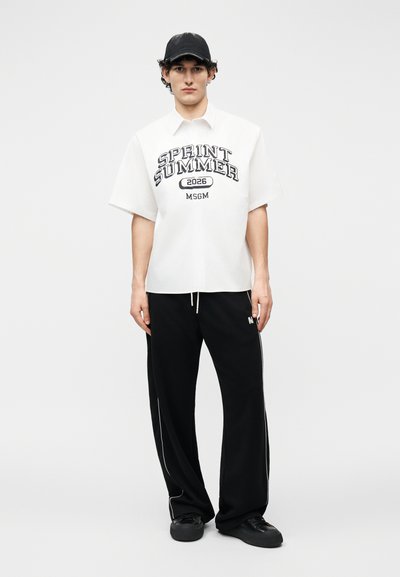 Jeune homme portant une chemise blanche à manches courtes avec le texte "SPRINT SUMMER 2026 MSGM", un pantalon noir, des baskets noires et une casquette noire.