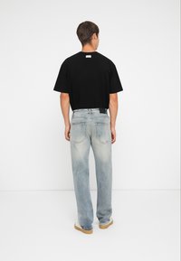 Zwarte katoenen T-shirt met korte mouwen, gecombineerd met lichtblauwe, iets versleten denim jeans en tan schoenen, gezien vanaf de achterkant.