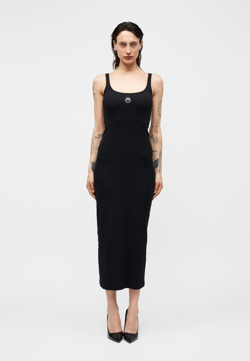 Γυναίκα με σκούρα μαλλιά, που φοράει μαύρο midi φόρεμα χωρίς μανίκια με κυκλικό λογότυπο στο στήθος και μαύρες μυτερές γόβες, στέκεται όρθια.