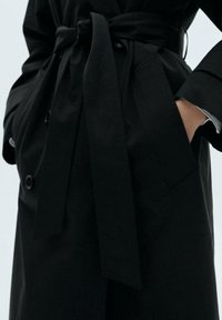 Schwarzer Trenchcoat mit einem Gürtel, verfügt über doppelte Knöpfe, tiefe Taschen und einen glatten, strukturierten Stoff. Klare Linien und modernes Design.