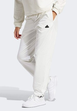Witte joggingsbroek van zachte stof met een elastische tailleband, zijzakken en een zwart Adidas-logo op de linkerdij. Draagt met witte sneakers.