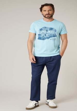 Homme debout, les mains dans les poches, portant un t-shirt graphique bleu clair, un pantalon bleu marine et des baskets blanches sur un fond uni.