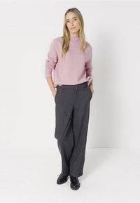 Růžový, ribovaný rolák v kombinaci s volnými tmavě šedými kalhotami. Modelka stojí se založenými rukama v kapsách a má na sobě černé boty.