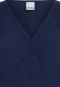 Haut bleu marine à col en V avec des détails à volants froncés le long du décolleté et du centre avant, avec une étiquette de marque blanche ICHI à l'intérieur du col.