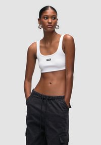 Top corto bianco a coste con spalline larghe e un'etichetta nera del brand. Il modello indossa pantaloni cargo neri con cordino, mostrando un look casual.