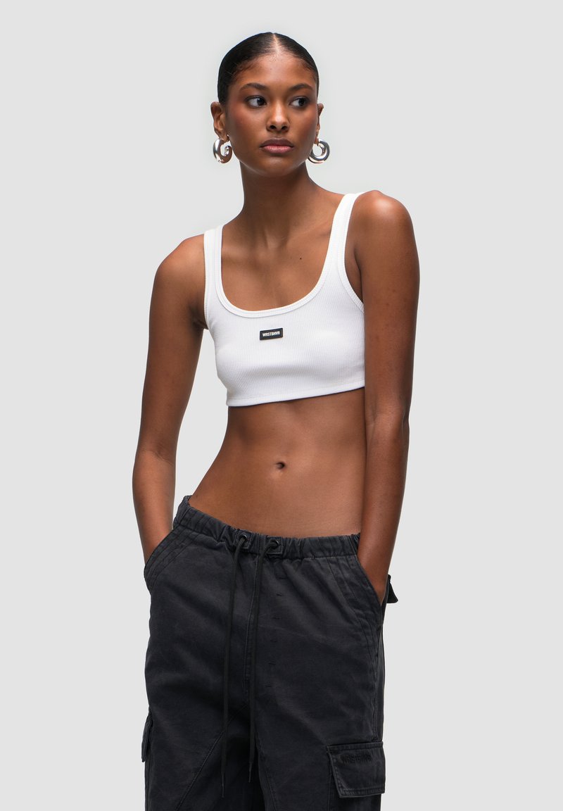 Top corto bianco a coste con spalline larghe e un'etichetta nera del brand. Il modello indossa pantaloni cargo neri con cordino, mostrando un look casual.