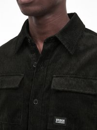 Close-up of a person wearing a black corduroy shirt with buttons, two chest pockets, and an "Urban Classics" label on the pocket.