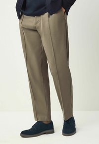 Pantalon beige avec plis sur le devant et poches latérales, associé à des chaussures en daim bleu foncé. Texture lisse avec une coupe ajustée et des lignes épurées.