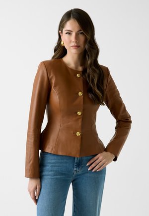 Femme aux longs cheveux bruns ondulés porte une veste en cuir marron cintrée avec des boutons dorés et des boucles d'oreilles dorées, assortie à un jean bleu sur fond blanc.