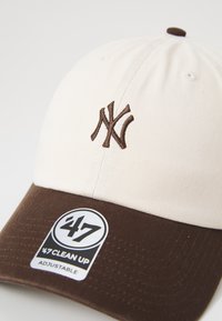 Pet met een crème kroon en een bruine rand. Heeft een donkerbruine geborduurde logo en een label met de tekst "47 CLEAN UP" in wit en zwart. Verstelbare pasvorm.