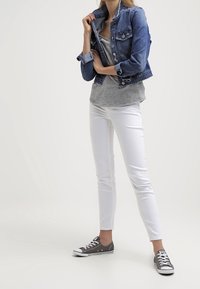 Žena upravující límec džínové bundy, oblečená v šedém tričku, bílých kalhotách a šedých teniskách, stojící na jednobarevném světlém pozadí.