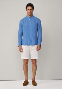 Camicia azzurra in lino a maniche lunghe con colletto alla cinese, frontale con bottoni e un piccolo logo sul petto, abbinata a pantaloni corti bianchi e scarpe marroni.