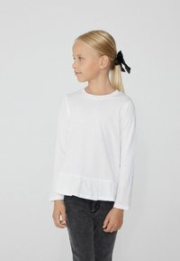 Biała koszula z długim rękawem i falbaniastym dołem, zestawiona z czarnymi dżinsami. Modelka ma proste blond włosy związane czarną kokardą.