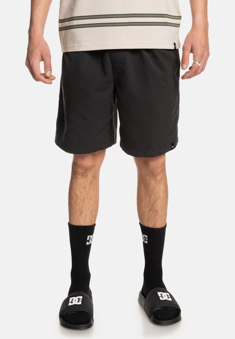 Black swim shorts made of lightweight fabric, featuring a flat waistband. Paired with black socks and slide sandals with logo accents.