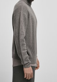 Maglione grigio a maglia con un collo alto, spalle scese e polsini a costine. Indossato sopra uno strato base più scuro. Tessuto morbido e texturizzato.