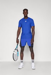 Modra športna majica z teksturnim vzorcem, v kombinaciji z modrimi in črnimi kratkimi hlačami. Model drži teniško lopar in nosi bele čevlje s črnimi črtami.