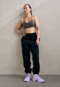 Atletická žena s opletenými vlasy, oblékající hnědou sportovní podprsenku Nike, černé joggingové kalhoty a světle fialové tenisky, stojící proti jedné obyčejné zdi.