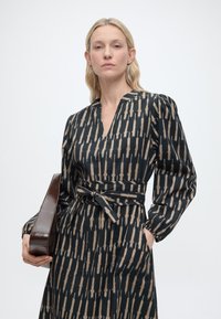 Γυναίκα με ξανθά μαλλιά που φοράει μαύρο φόρεμα με μπεζ κάθετο μοτίβο βουρτσιστών γραμμών, μακριά μανίκια και ζώνη στη μέση, κρατώντας καφέ τσάντα χειρός.