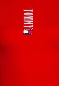 Red fitted t-shirt featuring vertical white and blue embroidered "TOMMY" with "TOMMY JEANS" underneath on a rectangular logo patch.