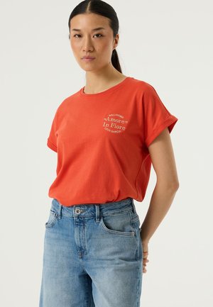 Maglietta rossa in cotone con maniche arrotolate e scollo rotondo. Presenta una scritta bianca sul lato sinistro del petto. Abbinata a jeans azzurri.