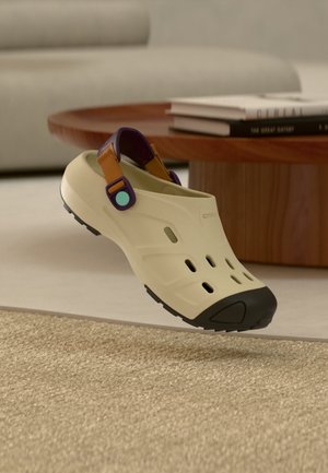 Μπεζ παπούτσι τύπου clog που φοριέται χωρίς δέσιμο με μαύρο προστατευτικό μύτης, μοβ και καφέ ρυθμιζόμενο λουράκι στη φτέρνα, τοποθετημένο κοντά σε στρογγυλό ξύλινο τραπέζι με στοιβαγμένα βιβλία.