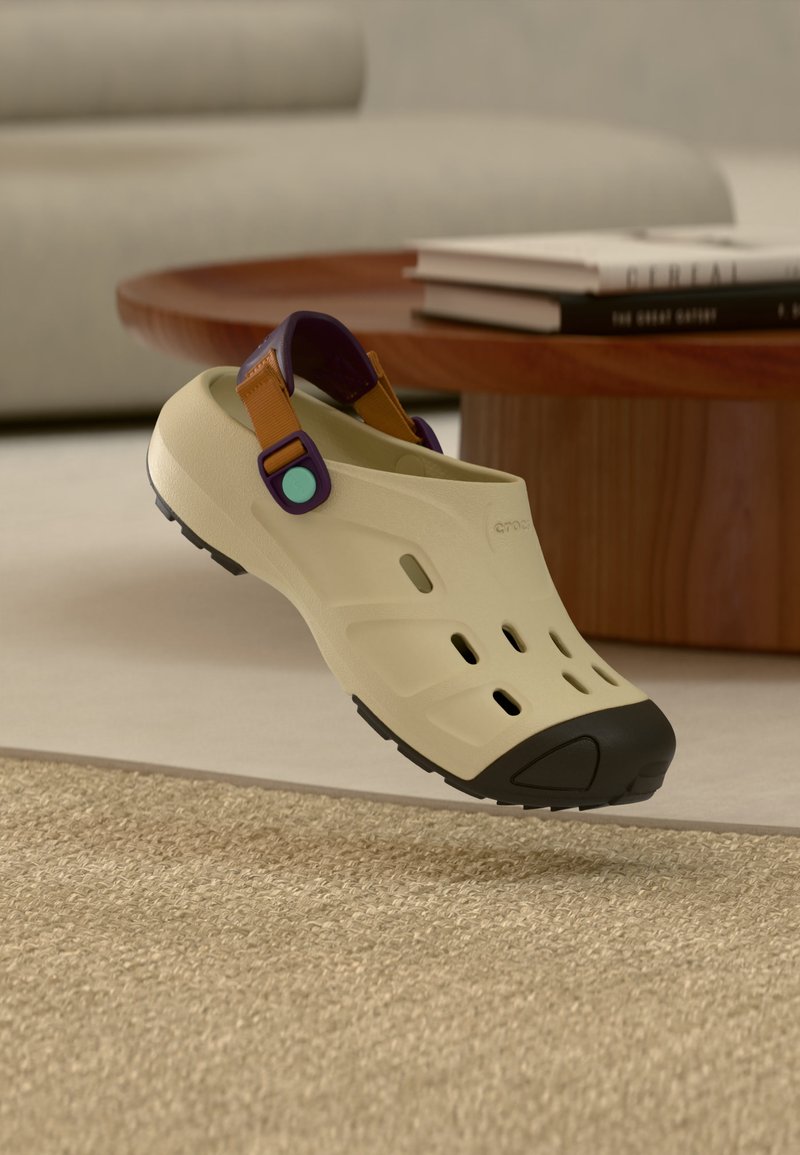 Sabot beige à enfiler avec embout noir, bride de talon réglable violet et marron, reposant près d'une table ronde en bois avec des livres empilés.