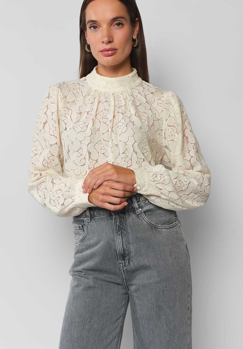 Cremefarbene Spitzenbluse mit hohem Hals, Puffärmeln, floralem Muster und knöpfbaren Manschetten, kombiniert mit grau hochtaillierten Jeans.