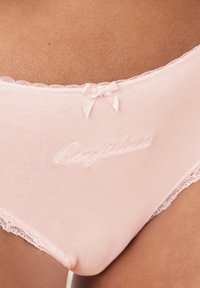 Culotte en coton rose clair avec une bordure en dentelle. Présente un texte brodé "Confidense" et un petit accent en forme de nœud sur la taille.
