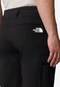 Black cargo pants with a smooth texture, featuring a back pocket and a white logo on the right side. Minimalist design with practical details.