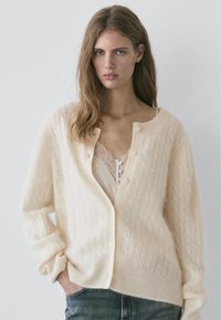 Roomkleurige kabelgebreide cardigan met knoopsluiting aan de voorkant, geribbelde manchetten en zoom. Draagt over een top met kanten randje, gecombineerd met spijkerbroek.